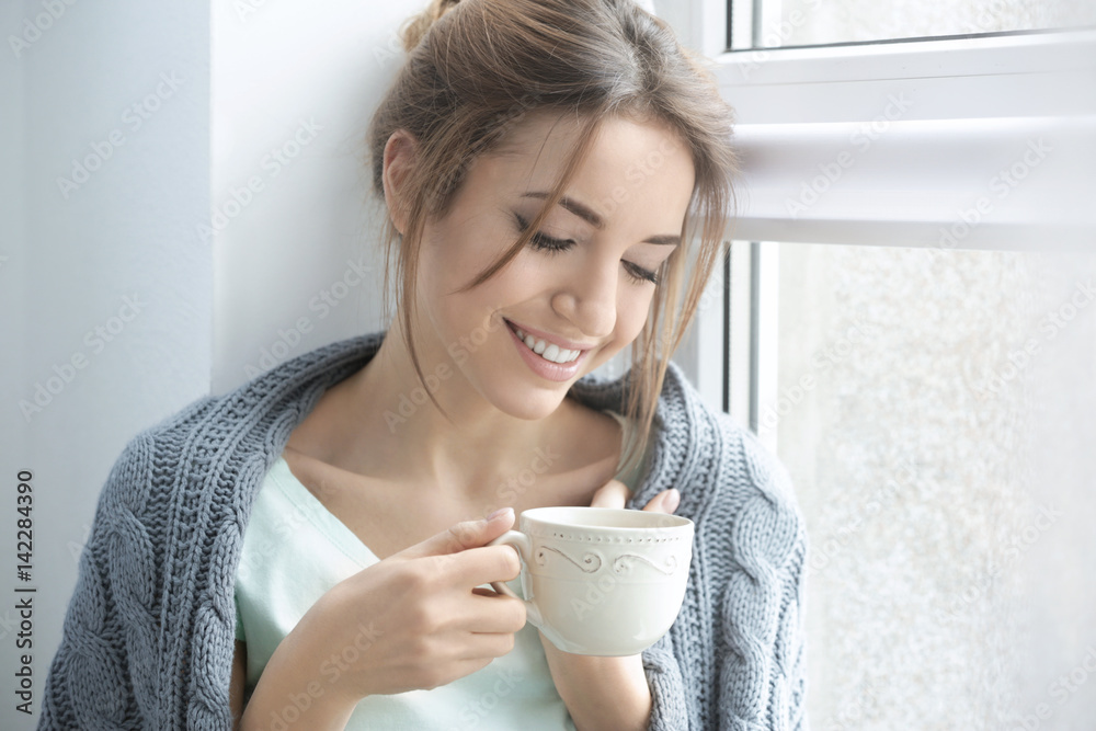 Beautiful young woman drinking tea near window at home