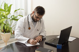 Fototapeta  - Lekarz w białym kitlu w gabinecie lekarskim siedzi przy stole i wypełnia dokumenty. Lekarz podczas pracy. 