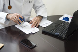 Fototapeta  - Lekarz w białym kitlu siedzi przy stole i przystawia pieczątkę do dokumentu. Ręce lekarza przystawiającego pieczątkę na recepcie. Lekarz wypisuje receptę przy biurku.