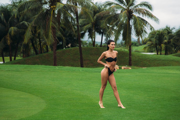 Wall Mural - Young sexy and beautiful brunette fitness girl in black swimsuit posing turning booty shows ass at the green grass of Golf club. Sexy model portrait with perfect body. Concept of summer holiday