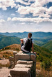 © Oksana - silhouette of man with backpack sitting with his back to the photographer on the big rock and looking at the panorama of mountains , sky and clouds