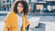 © skyNext - Portrait of young beautiful curly Brazilian woman with perfect smile in stylish yellow coat while sitting on a concrete bench with copy space for young advertising text message or promotional content
