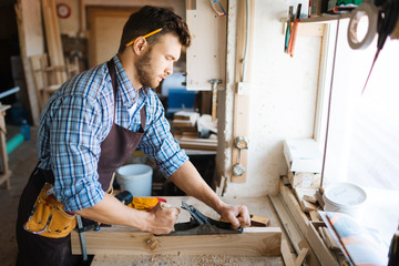 Wall Mural - Skillful bearded woodworker in checked shirt smoothing plank with jointer plane, shavings scattered on table