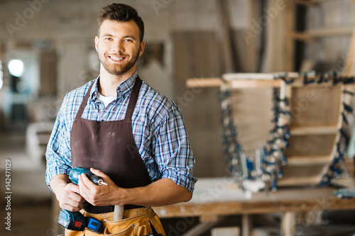 Cuadros en Lienzo Waist-up portrait of smiling bearded craftsman with electric drill in hands stan