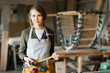 Waist-up portrait of confident fair-haired woodworker with tape measure in hands posing for photography while standing on background of workshop