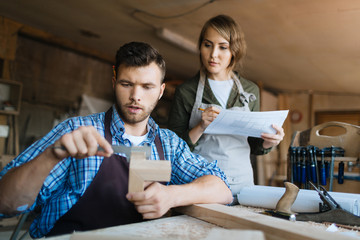 Wall Mural - Handsome bearded craftsman measuring width of wooden piece with vernier caliper while his pretty female apprentice taking notes in workshop