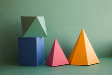 Abstract Geometrical Solid Figures Still Life. Colorful Three-dimensional Pyramid Prism Rectangular Cube Arranged On Green Background. Yellow Blue Pink Malachite Colored Objects Textured Paper Surface