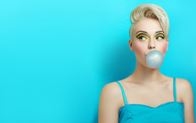 Fashionable Girl With A Stylish Haircut Inflates A Chewing Gum. The Girl In The Studio On A Blue Background. The Girl's Face With Bright Makeup And Yellow With Black Shadows On The Eyes.