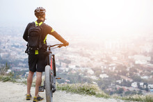 Unrecognizable European Male Cyclist Relaxing On Top Of Mountain, Keeping Hands On Handlebar Of His Electric Pedal-assist Bicycle, Enjoying Amazing Urban Landscape Below. Travel And Active Lifestyle