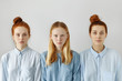 © WHstudio Leushin N - Studio shot of three college girls or sisters dressed in shirts posing against white wall background having neutral face expressions. Blond girl standing between two red haired female friends