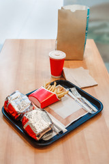 Closeup shot image of two burgers wrapped in tinfoil, french fries potato chips, red cup of coffee or tea on tray on wooden table in fast food restaurant