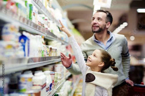 Cuadros en Lienzo Happy girl pointing at pack of youghurt on upper shelf in supermarket