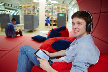 Portrait of smiling young man working in creative office workspace, relaxing on bean bags while using digital tablet and headphones