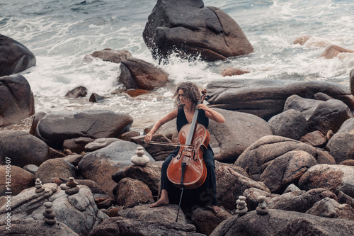 Plakat piękna młoda kobieta gra na wiolonczeli na kamienistej plaży w burzliwej pogody