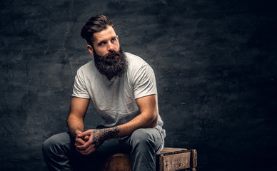 Bearded male with tattoo on arm dressed in a white t shirt sits on a wooden box.