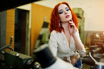 Wall Mural - Red haired girl wear on short denim shorts and white blouse posed at industrial machine at the factory.