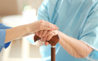 Wall Mural - Young doctor holding hands of elderly woman on light background, closeup