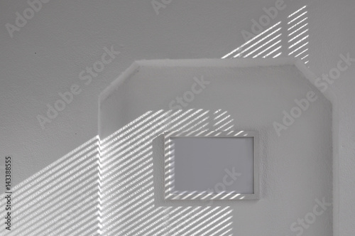 Fototapeta na szkło Tło architektoniczne. Biała otynkowana ściana z ukośnym cieniem od żaluzji i biała pusta rama.