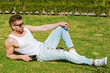 © T.Den_Team - A young sexy european and american man with biceps and a good figure with bristles lies on the green grass in a park in the city in the sunlight with a mobile phone  and blue jeans and goggles