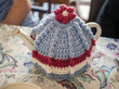 Vintage style teapot and knitted wool tea cosy or cozy on a patterned table cloth with a lace mat next to it.