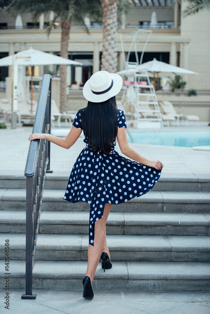 A girl with long straight hair in a blue dress with polka dots, a white hat  and black high-heeled shoes walking up the stairs Stock Photo