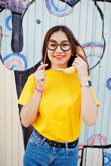 Beautiful fun teenage girl with banana at hand, wear yellow t-shirt, and fake glasses near graffiti wall. Speaking on phone.