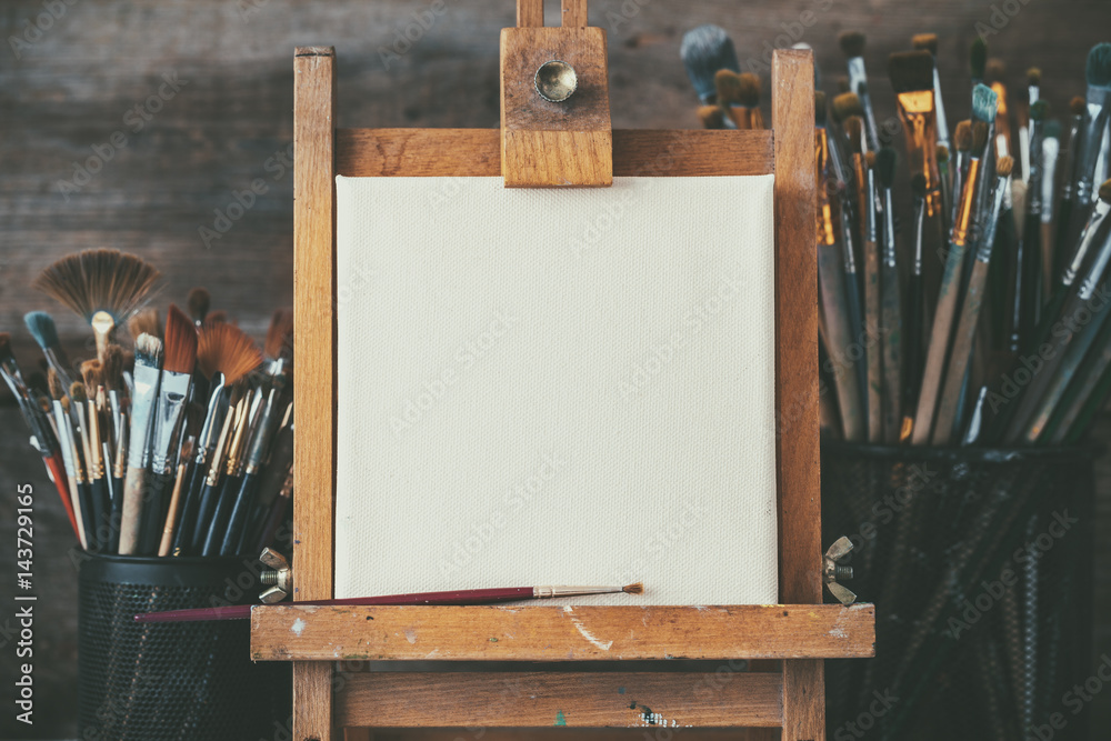 Artistic equipment in a artist studio: empty artist canvas on wooden easel and paint brushes Retro toned photo. - obrazy, fototapety, plakaty 