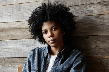 Indoor shot of stylish young Afro American female with curly hair wearing trendy denim clothing looking at camera with neutral calm expression, sitting on chair at coffee shop, waiting for friends