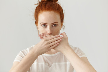 Portrait of serious young ginger European woman covering mouth with both hands keeping a secret. Freckled redhead female in blouse doesn't want to spread rumors or some confidential information