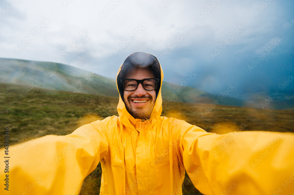 Selfie portrait of a smiling and laughing traveller man in yellow raincoat and glasses in the clouds mountains in stromy weather with rain - obrazy, fototapety, plakaty 