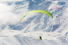 Parachute Sky-diver Flying In Clouds Above Mountains With Fresh Snow On Sunny Winter Day In The Ski Resort. Travel Adventure Concept. Space For Text