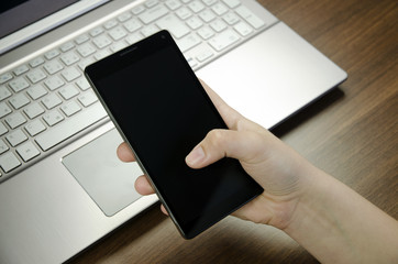 Poster - Closeup view of a woman's hand with fingers holding a black smartphone on silver laptop keyboard lying on a wooden mat blurring