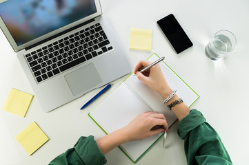 Working at desk with notepad and laptop computer. Top view of female person hands writing in note book sitting at white table with smartphone, portable computer and glass of water