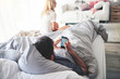 © Connect Images - Man in bed using smartphone, woman sitting at end of bed