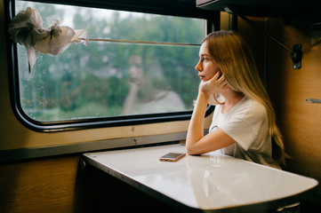 Very beautiful and attractive skinny teen girl with long hair in sleeveless shirt traveling by train alone on vacation and looking at window to summer landscape. Deep mood concept.