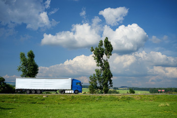 Blue truck driving on the road next to tall trees in a rural landscape under a blue sky with dramatic white clouds