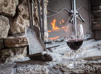 horizontal image of a glass of red wine sitting on the hearth with the fire burning in an old rustic stone fire place.