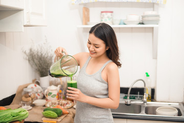 Wall Mural - Smiling asian woman making smoothie with fresh vegetables in the blender in kitchen at home.