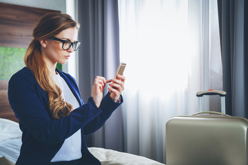 Wall Mural - Business woman with suitcase in modern hotel room using laptop and smartphone