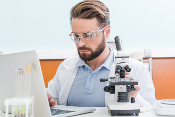  concentrated bearded scientist working with microscope and laptop in laboratory