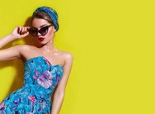 A Beautiful Young Tanned Girl With A Headscarf On Her Head And Wearing Sunglasses Stands Near A Yellow Wall In The South. Fashionable Blue Summer Dress, Bright Make-up, Tan.