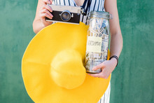 Woman Holding A Jar Full Of Money Savings For Summer Vacation With Yellow Hat And Photo Camera On The Green Background