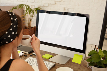 Cropped rear view of successful businesswoman busy using computer at the office desk, mock up. Cropped view of young female manager working in office drinking coffee, PC with blank screen copy space