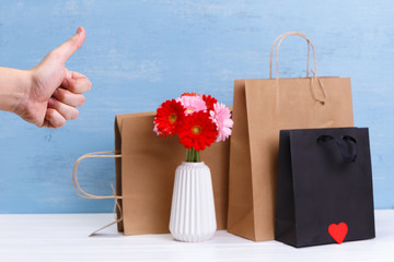 Mockup of blank shopping bags. Gerbera flowers and red heart. Hand with thumb up. Concept for sales or discounts. Blue wooden rustic board.