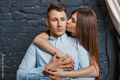 Beautiful young girl kisses a guy on the cheek near a brick wall ...