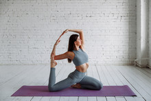 Young Attractive Smiling Woman Practicing Yoga, Sitting In One Legged King Pigeon Exercise, Eka Pada Rajakapotasana Pose, Working Out, Wearing Sportswear, Grey Pants, Bra, Indoor Full Length, Home