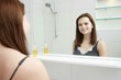 © Saksoni - morning and people concept - young woman looking to mirror at home bathroom