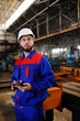 © Evgeniy Kalinovskiy - A man in a white helmet and working construction clothing with a computer tablet in his hands against the backdrop of a factory, machines, production