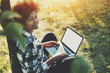 © skyNext - Attractive black teen girl in checkered shirt with laptop sitting on grass of spring meadow on sunny summer day, charming mixed female with curly afro hair and net-book leaning on apple tree in park