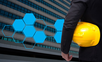 Wall Mural - back view of smart businessman in black suit with yellow safety helmet on building background and blank hexagon shaped pattern, color tone effect, copy space.